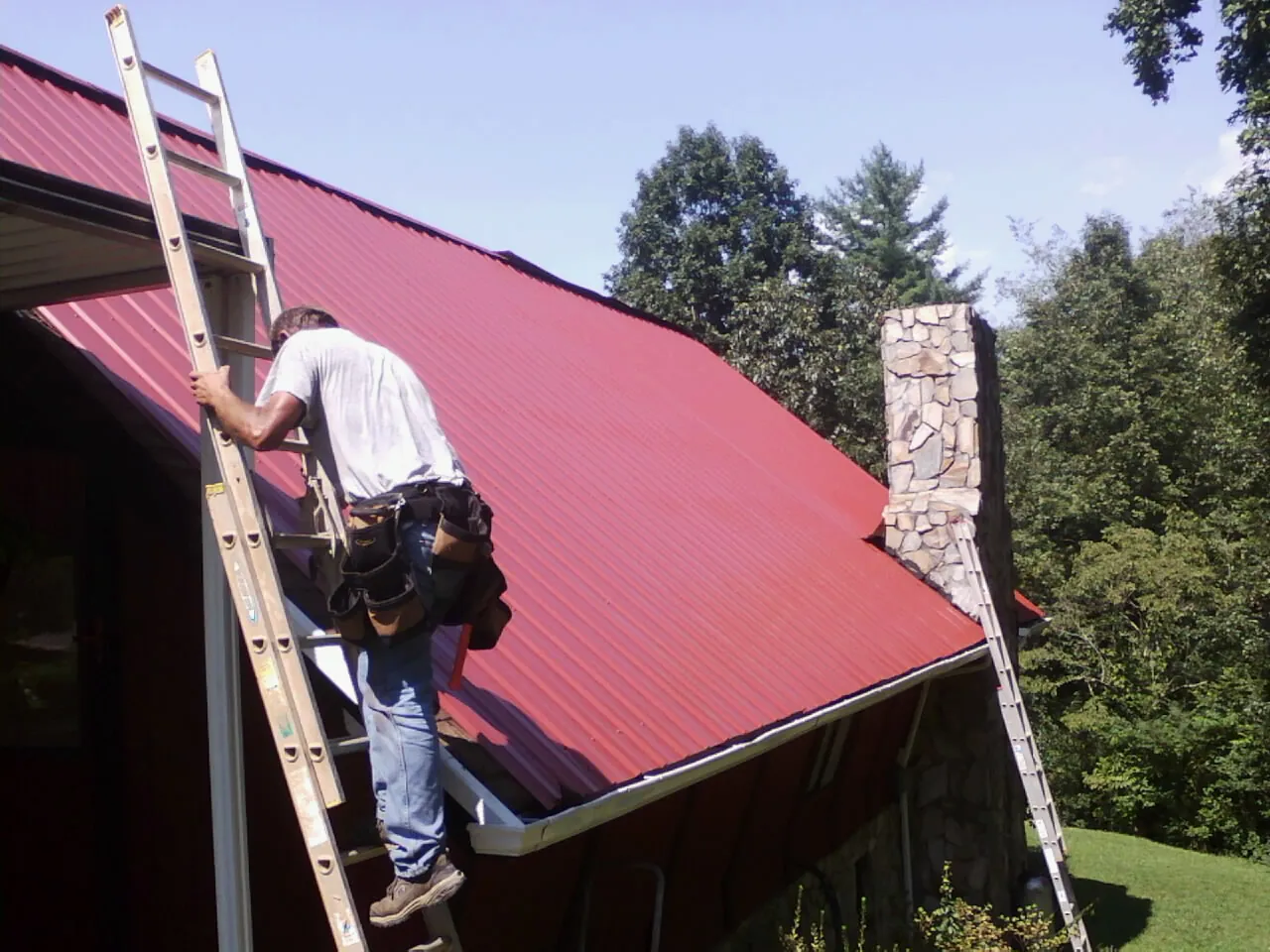 Roofer on a ladder beside a red metal roof and stone chimney — Soffit & Fascia Repair by Roofing Brothers in Rockwell